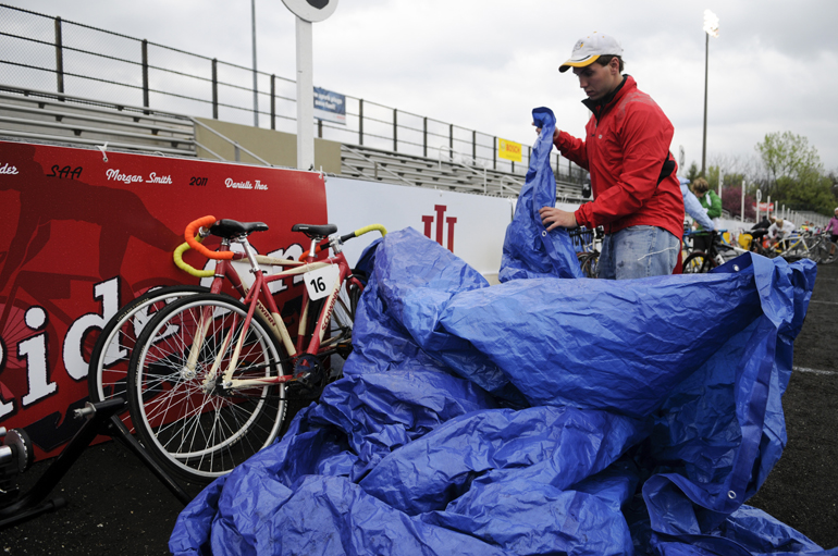 Ride On coach Isaac Simonelli takes the tarp off of his team’s bikes before the women’s Little 500 on Friday at Bill Armstrong Stadium. The IU Student Foundation put out the bikes early in the morning, but because of the pre-race rain, tarps were put out later. (Alex Farris | Indiana Daily Student)