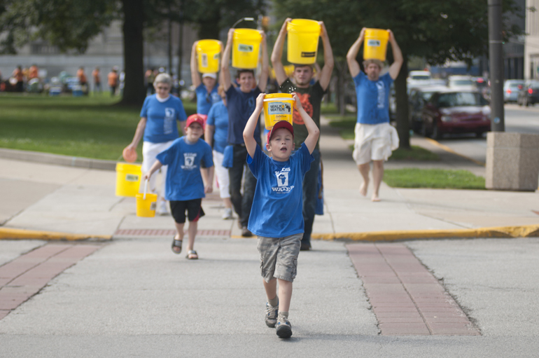 Jameson Chapman leads walkers across Michigan Street while carrying canal water during the third annual Walk for Water event downtown, Saturday, Aug. 2, 2014. The event raised money for a community clean-water well in Kager, Kenya. Attendees walked from American Legion Mall to the canal, filled buckets with water and walked back to the mall, simulating the path villagers without clean water have to take every day.